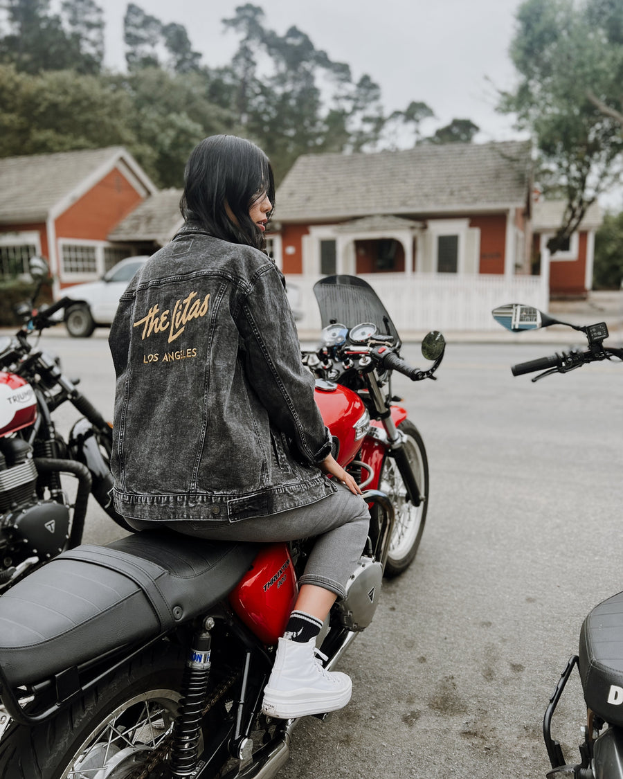 Person sitting on a motorcycle wearing a custom denim jacket with 'The Litas' logo, with a rural setting in the background.